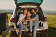 © Stockphotodirectors - A family happily shares a picnic during sunset, nestled in the trunk of their car. Green hills and a colorful sky create a beautiful backdrop for their joyous moment.