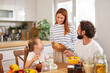 © Stockphotodirectors - A cheerful family shares breakfast in a bright kitchen, with a mother bringing a basket of pastries to the table, while a father and daughter enjoy their meal.