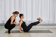 © TRAVELARIUM - Two adult women in black workout attire engage in pilates on mat. One assists other, ensuring proper form and technique. Setting is well-lit studio with white curtains, creating focused atmosphere