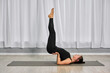 © TRAVELARIUM - Young adult female with light skin performs shoulder stand yoga pose on mat. Minimalist studio is styled with white curtains and light wood flooring, creating serene atmosphere