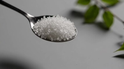 Aufkleber - Teaspoon full of granulated white sugar crystals, presented on a white background with green leaves in the background, suggesting a focus on sugar consumption and its alternatives