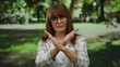 © Krakenimages.com - Senior hispanic woman with glasses stands in a park crossing arms to form an x against a vibrant green outdoor background.