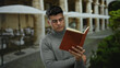 © Krakenimages.com - Young man in glasses reads a book on a terrace at an outdoor city restaurant, surrounded by greenery and umbrellas, providing a serene and studious ambiance.