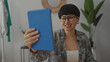 © Krakenimages.com - Woman smiling during a videoconference in a modern living room interior, holding a blue tablet, wearing glasses and casual attire, surrounded by contemporary home decor and plants.
