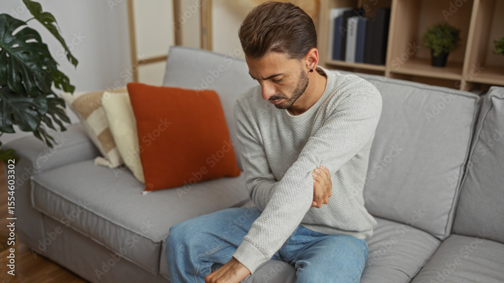 Young man sitting on a sofa in a modern living room massaging his sore arm, suggesting discomfort, home leisure, and contemporary interior design.