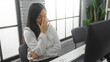© Krakenimages.com - Woman in office appearing tired with closed eyes and hand on face in a modern workplace setting, featuring natural light through large windows and indoor plants.