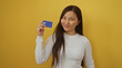 © Krakenimages.com - Woman smiling holding credit card against vibrant yellow background with long hair wearing white sweater asian female confident pose financial security concept