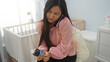 © Krakenimages.com - Woman multitasking, young and asian, sits in a baby room, balancing a phone call while examining a medicine bottle, highlighting the challenges of modern motherhood at home.