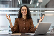 © Liubomir - A smiling woman in a brown shirt meditates with her hands in a mudra at her desk in a modern office setting.