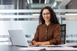 © Liubomir - A smiling woman works on a laptop in a modern office environment, radiating professionalism and positivity.