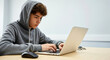 © DP - Young male student wearing a gray hoodie is focused on typing on a laptop at a wooden desk, showcasing concentration and engagement in digital learning activities