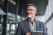 © cut - Happy middle aged business man ceo wearing suit standing in office using digital tablet. Smiling mature businessman professional executive manager looking away thinking using modern technology.