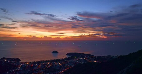 Wall Mural - A breathtaking sunset paints the sky in vivid hues of orange and blue over a coastal town, with lush green hills framing the scene. A distant hilltop structure adds a serene tranquil landscape.