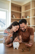 © Migma_Agency - Happy couple saving money together at home putting coins in piggy bank