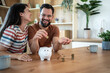 © Migma_Agency - Happy couple saving money together at home putting coins in piggy bank