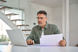 © insta_photos - Serious busy middle aged mature man wearing eyeglasses holding paper bill checking insurance invoice or tax document calculating expenses working online at home using laptop computer.