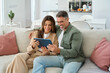 © insta_photos - Candid photo of happy middle aged couple using digital tablet relaxing on couch at home. Smiling mature man and woman holding tab browsing internet on pad device sitting on sofa in living room.