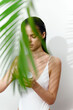 © VICHIZH - Beautiful woman applying skincare product in a white swimsuit with palm leaves in the foreground, representing natural beauty and self-care.