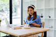 © David - Smiling girl student wear headphone study online with teacher, happy asian woman learn language listen lecture watch webinar write notes look at laptop sit in cafe, distant education