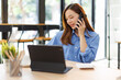 © David - Smiling asian woman writing notes while making phone call and using laptop at workplace.