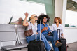 © Koldo_Studio - Group of multi ethnic friends waving goodbye while sitting and chatting in the waiting area of an airport terminal with luggage