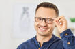 © Studio Romantic - Portrait close up shot of bearded smiling Caucasian man trying on new glasses in ophthalmology clinic or optical store with eye chart on background. Young man choosing eyeglasses to improve vision.