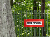 Czerwony znak „Droga pożarowa” na tle zielonego lasu, umieszczony na ogrodzeniu. Ostrzeżenie o zakazie wstępu w przypadku zagrożenia pożarowego lub akcji ratowniczej