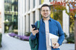 © Liubomir - A smiling man holds a phone and laptop outside a modern building on a sunny day. He wears glasses and a backpack.