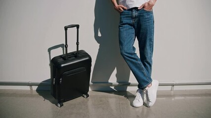 Poster - Young male tourist wearing jeans and white sneakers is leaning against a white wall with his black carry on suitcase next to him, waiting at the airport for his flight