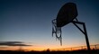 © Madasukma - A basketball hoop stands silhouetted against a gradient sunset sky, showing a weathered net and a serene, peaceful atmosphere.