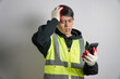 © yashis007 - A frustrated Asian man in a high-visibility safety vest and work gloves holds his phone while clutching his head, indicating stress or a workplace issue.