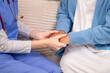 © Rakchanok - Young female nurse holding hands with senior woman, both smiling with trust and warmth during health support session in cozy home setting, healthcare, caregiver, take care, consult.