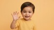 © Ardi - Smiling toddler waving hello with an open hand, set against a solid light orange background.