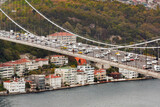 The Bosphorus Bridge, spans a body of water, packed with vehicles under an overcast sky in Istanbul. Residential buildings line the waterfront below, while a lush, green hill rises in the background.