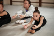 © DragonImages - Caucasian teenage girl stretching on floor during rhythmic gymnastics training with Caucasian young adult woman instructor assisting, both reaching forward with arms extended in gym setting