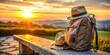 © Roman - A well-worn backpack sits on a bench next to two hats, soaking in the beautiful sunset. The sky glows with warm colors as nature envelops the peaceful landscape in the background