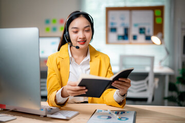 Poster - Young Asian businesswoman wearing headset is reading notes and working with desktop computer in modern office