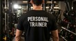 © ARONNABOSU - Personal Trainer at Gym: A personal trainer stands in a gym, his back to the camera, showcasing his 'PERSONAL TRAINER' shirt. The background features various gym equipment.