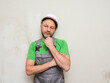 © mark_gusev - A workman in a green shirt and gray overalls hands crossed, looks tired. Portrait of a male general laborer on a construction site in white safety helmet. Model in his 40s with grey beard.