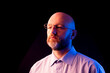 © mark_gusev - A man with a beard and glasses is standing in front of a black background. Male model in light shirt, bald head illuminated with orange and blue light. Office worker studio shot.