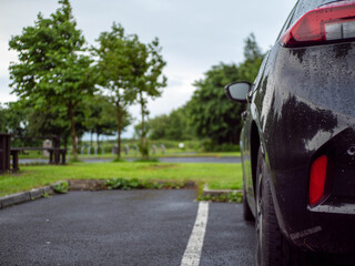Wall Mural - Black family car parked in a parking lot of a park with barbecue or dining area, selective focus. Nobody. Car surface is with rain drops. Outdoor activity. Travel and tourism.