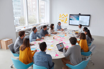 Poster - Diverse team collaborating in a modern office. Brainstorming session around a table with tech, notes, and digital display. Perfect for teamwork, innovation, and growth.