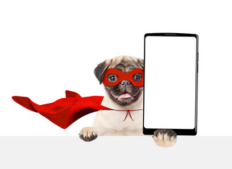 Poster - Happy Pug puppy wearing superhero costume shows big smartphone with white blank screen in it paw above empty white banner. Isolated on white background