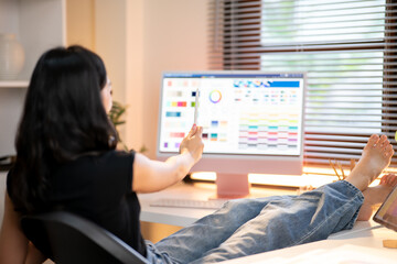 Fototapete - Asian woman designer with stylus looking at color chart on computer sitting legs up on working table