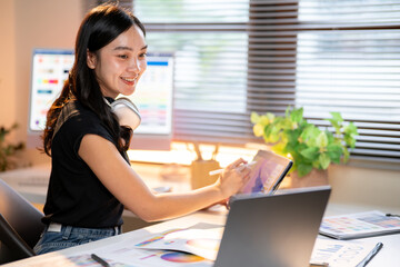 Fototapete - Asian woman designer drawing on tablet looking at laptop as sits with color chart at office's table.