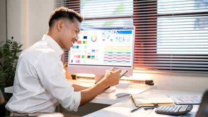 Fototapete - Happy asian man designer looking at color chart on tablet and sitting with computer on working table