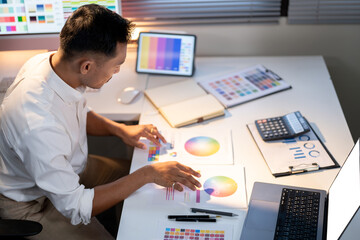 Fototapete - Asian man designer looking at color palettes while sitting with laptop and tablet on working table.