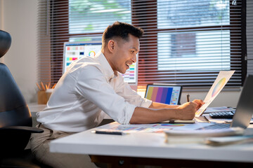 Fototapete - Happy asian man designer looking at color chart writing in book aside tablet on office working table