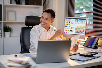 Fototapete - Smiling asian man designer holding pen looking away from color chart sits at office's working table.