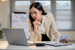 © bongkarn - Close up businesswoman or office worker leaning over table holding pen under chin looking at laptop.